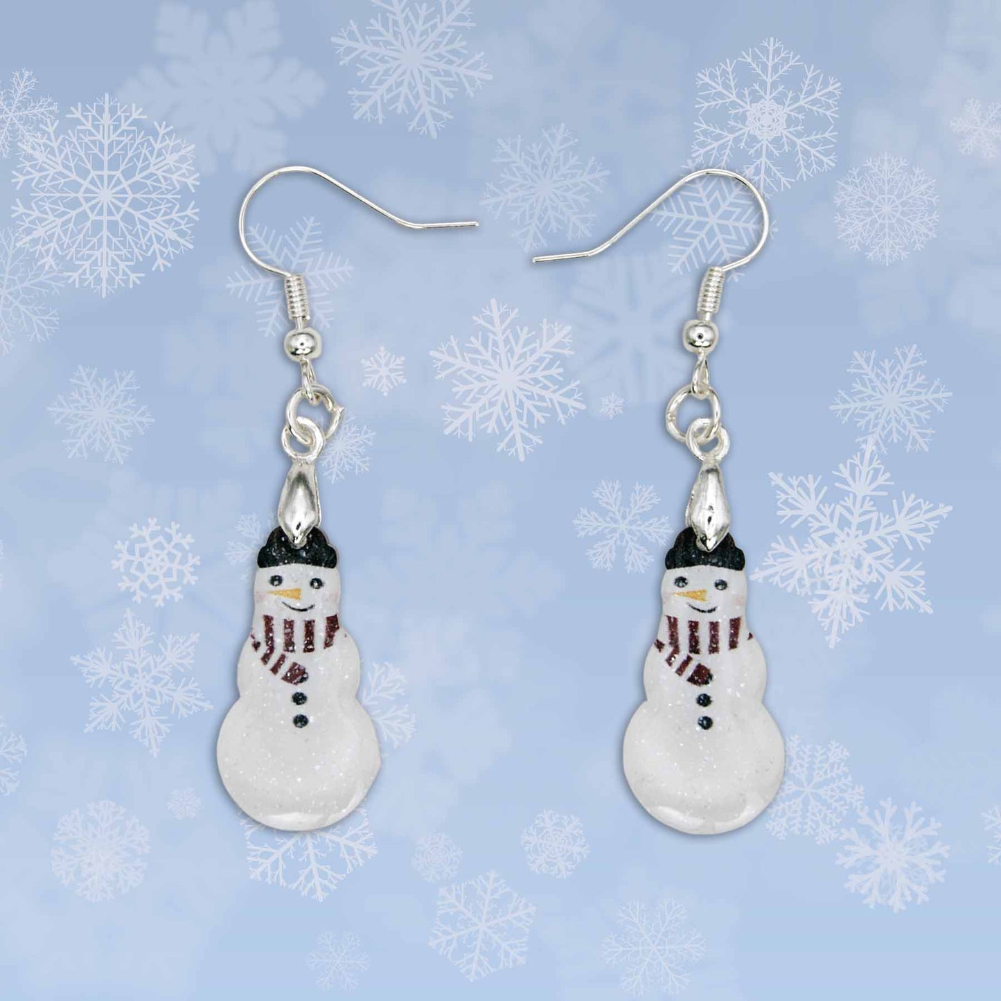 Dangling snowman earrings with silver hooks, glittery white bodies, black hats, red and black striped scarves, and black button details. The snowmen have orange and red facial features. They are displayed against a blue background with white snowflakes.