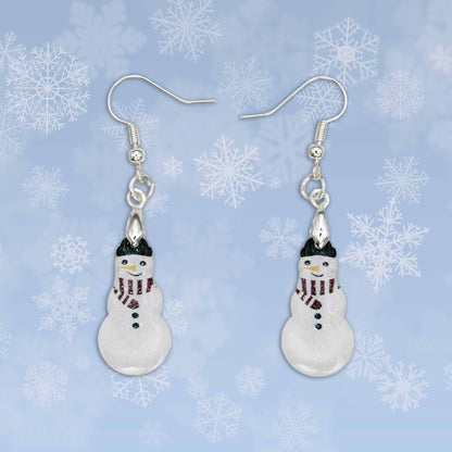 Dangling snowman earrings with silver hooks, glittery white bodies, black hats, red and black striped scarves, and black button details. The snowmen have orange and red facial features. They are displayed against a blue background with white snowflakes.